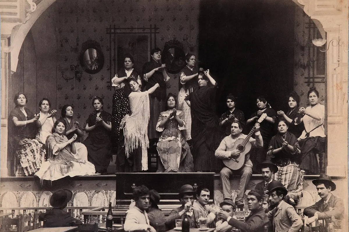 A black and white photograph of a café cantante in Seville around 1888, with two women dancing and others clapping on a raised stage, a guitarist seated at the right and men at tables in front