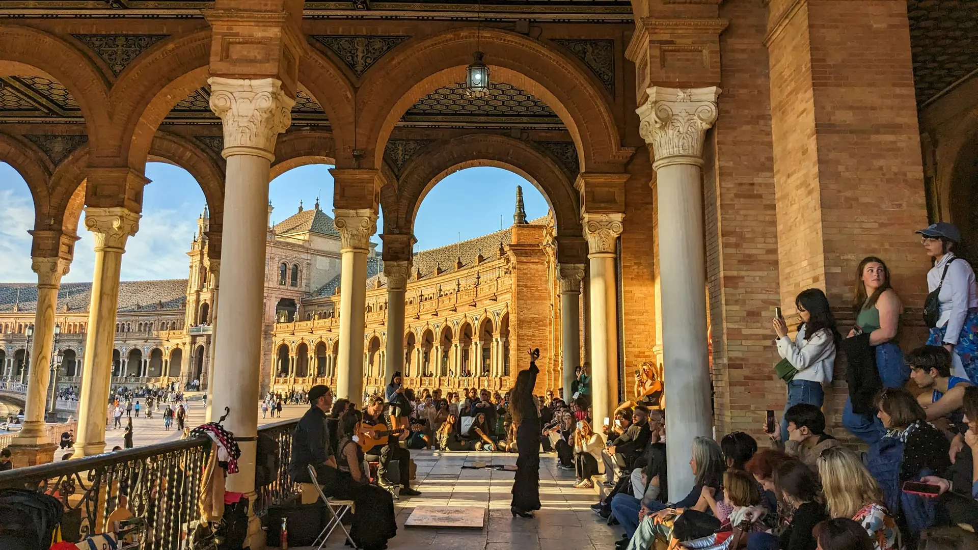 A flamenco dancer in black raises one arm in the golden light under the brick arcades of Plaza de España in Seville, with a guitarist beside her and a crowd seated around her